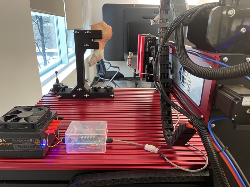 On the CNC bed: the limb model on its fixture, a joystick, and a power supply beside a small clear enclosure.